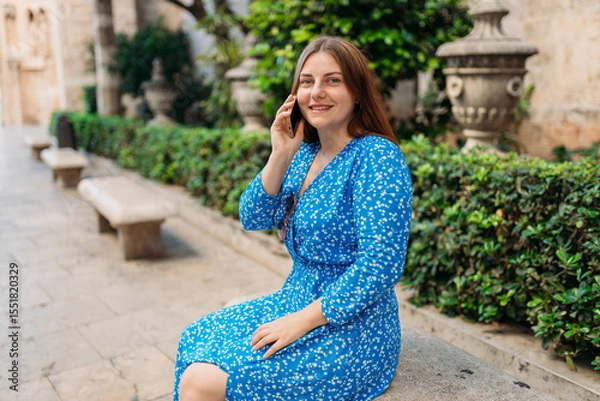 Obraz Young happy redhead 30s woman making phone call over mobile phone in the city. Middle age woman talking on smartphone on urban background. Traveling Europe in summer