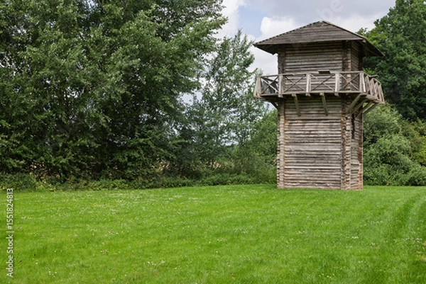Obraz A reconstructed Roman watchtower stands tall in a lush green field, surrounded by dense trees under a bright sky, evoking ancient history in Utrecht
