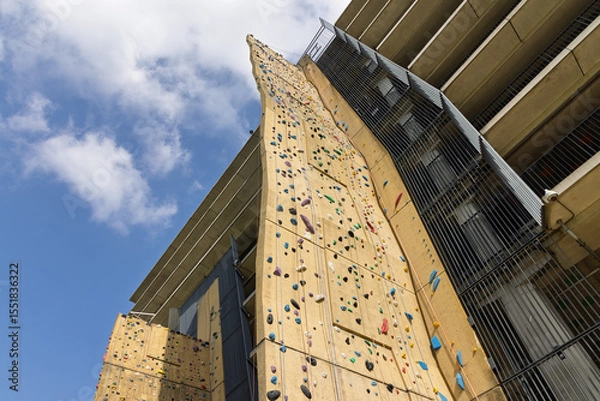 Obraz A massive outdoor climbing wall, studded with colorful holds, stretches skyward beside a modern building, offering an impressive challenge at Utrecht Science Park.