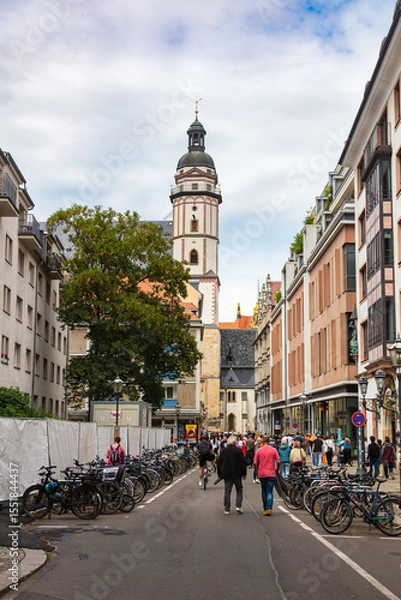 Obraz Leipzig, Germany – June 7, 2025:  St. Thomas Church (Thomaskirche) tower rising above a busy street filled with pedestrians and bicycles. church is associated with composer Johann Sebastian Bach.