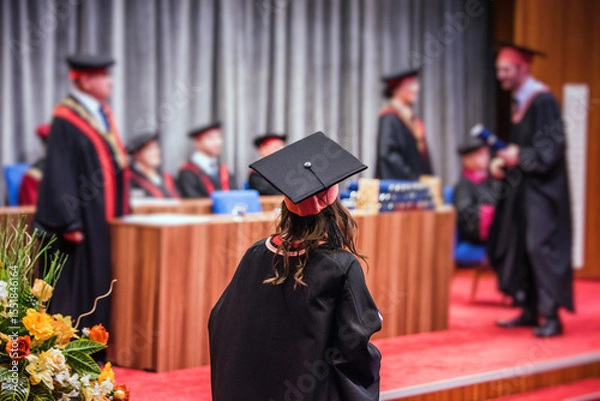 Fototapeta Back view of a female graduate walking on stage to receive her diploma during a graduation ceremony. She is wearing a traditional graduation gown and a square academic cap known as a mortarboard
