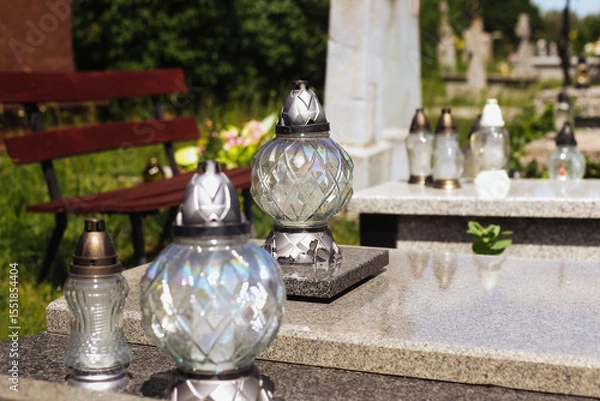 Fototapeta Grave with decorative glass lanterns. Cemetery memorial with candles on tombstone. Peaceful resting place. Daylight scene with greenery and bench. Symbol of memory and remembrance. Polish tradition.