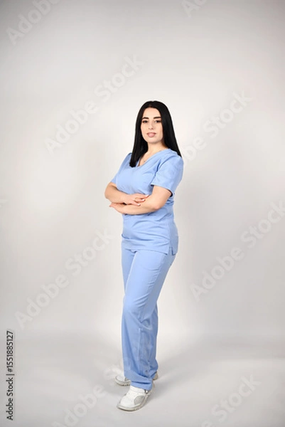 Fototapeta A cheerful, smiling female nurse in a full-length blue medical suit on a white background. Portrait of a young nurse. The image conveys professionalism and experience in the field of healthcare