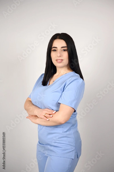 Fototapeta A beautiful, smiling female nurse in a blue medical suit on a white background. Portrait of a young nurse. The image conveys professionalism, confidence and experience in the field of healthcare
