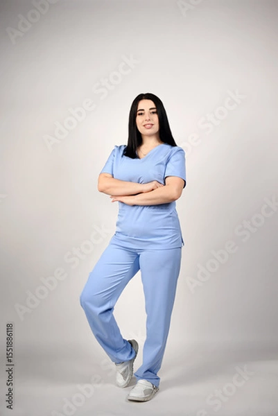 Fototapeta A beautiful, smiling female nurse in a full-length blue medical suit on a white background. Portrait of a doctor. The image conveys professionalism and experience in the field of healthcare