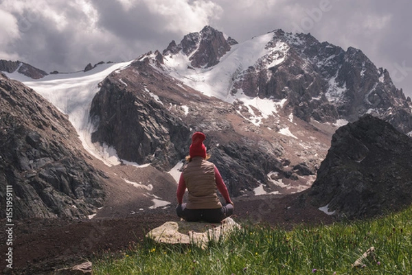 Obraz Rear view of a woman sitting cross-legged on a rock, wearing a red beanie and vest, meditating or resting while facing dramatic snow-covered mountain peaks under a cloudy sky. 
