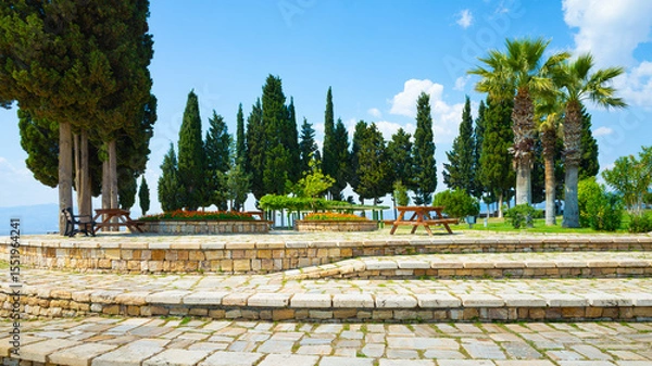 Obraz Public park with paved stone terraces, benches, picnic tables, and tall trees in Hierapolis near Pamukkale, Turkey. Sunny summer day with blue sky and scattered clouds in peaceful green surroundings.