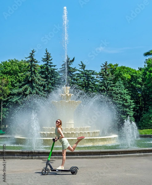 Obraz Happy young woman having fun with an electric scooter in a city park by a fountain on a sunny summer day.Urban mobility and summer lifestyle.Eco-friendly transport concept