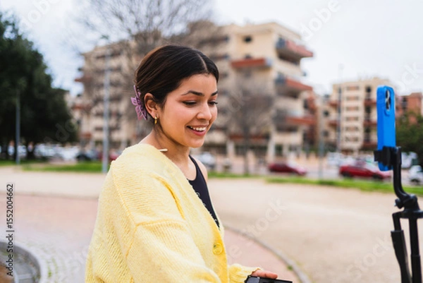 Fototapeta Young woman vlogger recording video blog on phone using tripod in city park