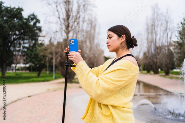 Fototapeta Young woman creating video content for social media using a smartphone on a tripod in a park