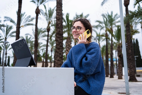Fototapeta Young woman attending online classes or smart working using digital tablet and smartphone outdoors in a park with palm trees
