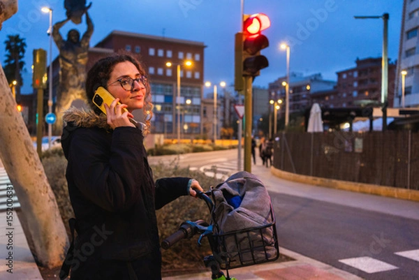 Fototapeta Young woman using mobile phone while waiting with her bicycle at red traffic light in urban environment at dusk