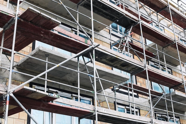 Fototapeta Building under renovation process. Metal scaffolding on construction site. Exterior wall insulation and repair. Urban maintenance in progress. Industrial framework structure in city renovation work.
