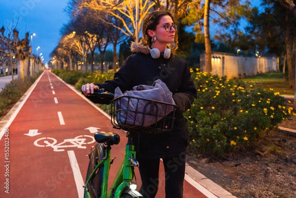 Fototapeta Student walking with her bicycle on a bike path at dusk, enjoying the fresh air and the urban landscape