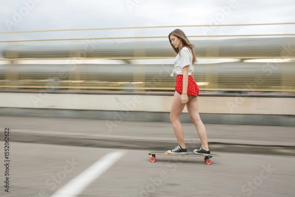 Fototapeta Young woman riding a longboard in motion on an urban street. Dynamic summer vibe with blurred background