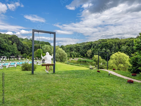 Obraz Swinging into Summer: Woman Enjoying a View of a Luxury Resort with a Pool and Forest,Rear View.Modern Recreation: Enjoying the Outdoors on a Stylish Swing at a Scenic Holiday Destination