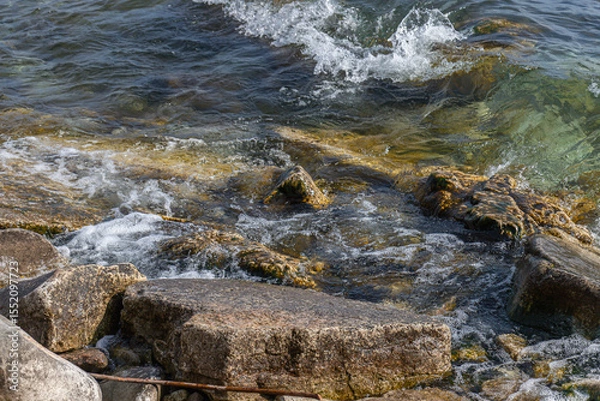 Fototapeta Clear lake water splashes over mossy rocks and stones on a rugged shoreline, capturing natural textures and motion.