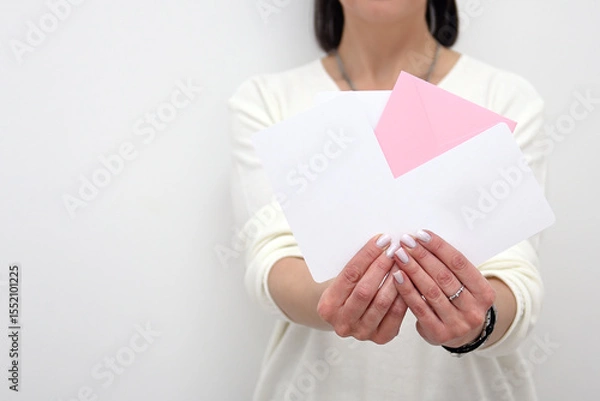 Fototapeta A girl in light-colored clothes holds several white and pink advertising envelopes in her hands. A flat envelope in a woman's hands with an empty space. Invitation, package, and letter
