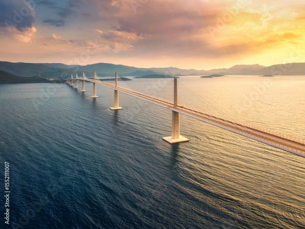 Obraz Aerial view of modern Peljesac Bridge and blue sea at colorful sunset in summer. Croatia. Top drone view of bridge during golden hour, mountains, reflection in water and orange sky. Architecture