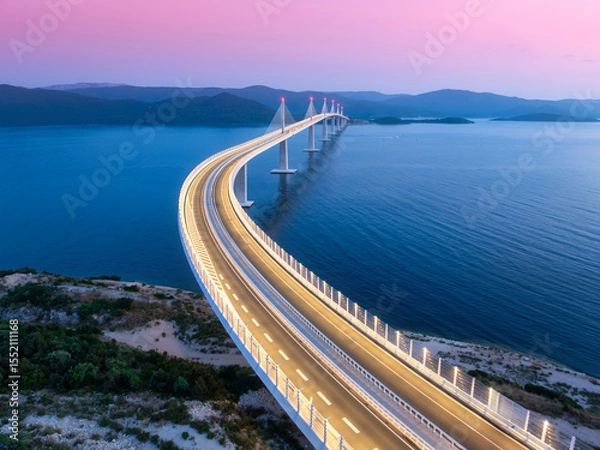 Obraz Aerial view of modern Peljesac bridge and blue sea at night. Croatia. Bridge with illumination, mountains, reflection and pink sky at sunset. Top drone panoramic view of road, street lights at dusk