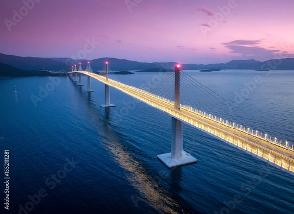 Obraz Aerial view of modern Peljesac bridge and blue sea at night. Croatia. Bridge with illumination, mountains, reflection and pink sky at sunset. Top drone panoramic view of road, street lights at dusk