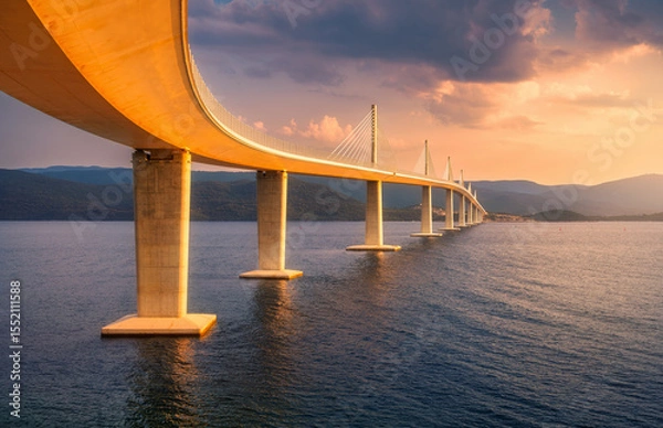 Obraz Modern Peljesac bridge and blue sea at colorful sunset in summer. Croatia. Beautiful Peljeski bridge during golden hour, mountains, reflection in water, orange sky with clouds. Architecture. Landscape
