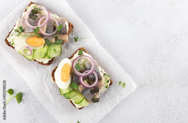 Fototapeta Traditional Danish smorrebrod with herring, egg, cucumber, onion and herbs on rye bread on white background. Copy space.