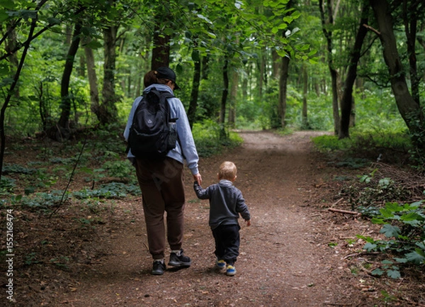 Obraz Nature walk with baby, family bonding in the forest. Mom holds baby's hand while walking along wide forest path