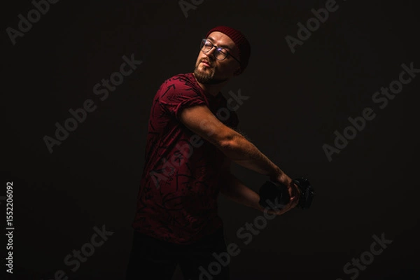 Fototapeta Stylish male photographer in red beanie and graphic tee posing in dramatic low key lighting, holding a camera against dark background. Urban creative vibe