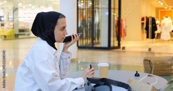 Obraz Young muslim woman wearing hijab is sitting at a table in a shopping mall, recording voice notes on her phone and taking notes on her tablet. Free and modern Muslim in city