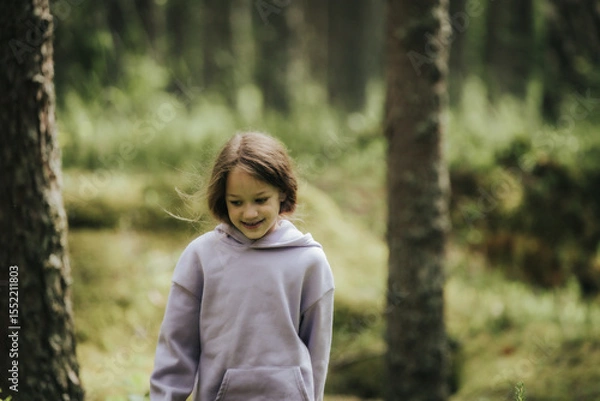 Fototapeta A young girl in a lavender hoodie and pink pants walks through a mossy forest, holding a plastic jar, possibly collecting insects or exploring nature.