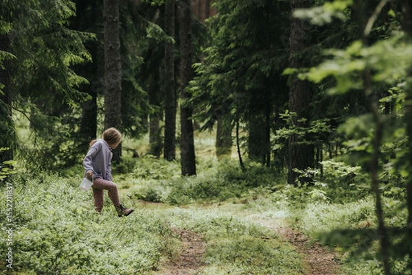 Fototapeta A young girl in a lavender hoodie and pink pants walks through a mossy forest, holding a plastic jar, possibly collecting insects or exploring nature.