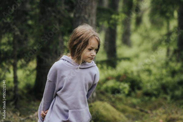 Fototapeta A young girl in a lavender hoodie and pink pants walks through a mossy forest, holding a plastic jar, possibly collecting insects or exploring nature.