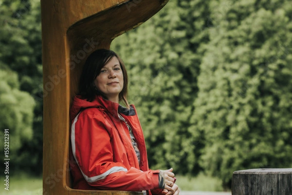 Fototapeta A woman in a red outdoor jacket leans against a large wooden structure with a calm expression, surrounded by a lush green forest background.