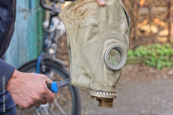 Fototapeta hands of one man holding one old gray army rubber gas mask with an knife in his head on an autumn street