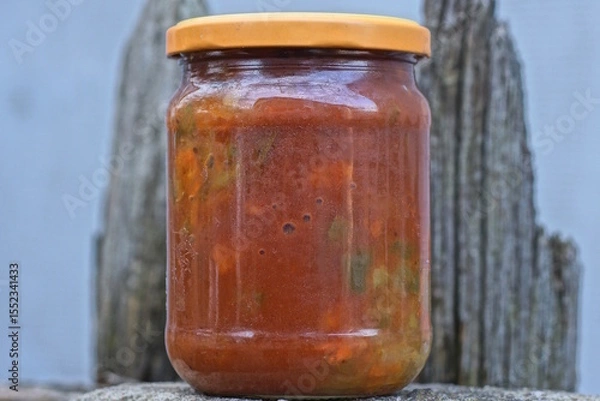 Fototapeta one closed glass jar with red lecho from canned pepper stands on a gray table on an autumn street