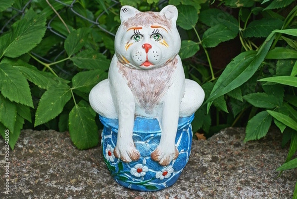 Fototapeta one large ceramic colored bank piggy bank with a figure of a cat stands on a gray table against the backdrop of green vegetation on a summer street