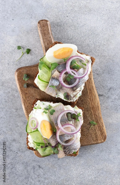 Fototapeta Danish smorrebrod with herring, egg, cucumber and red onion on rye bread on a wooden board. Top view