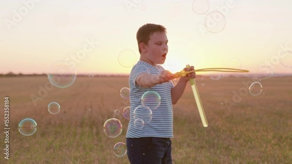 Obraz Child boy blows soap bubbles, having fun playing on field in sunset. Child boy plays with soap bubbles in park in sun nature. Happy childhood concept. Cheerful kid smiling, holiday in summer outdoors
