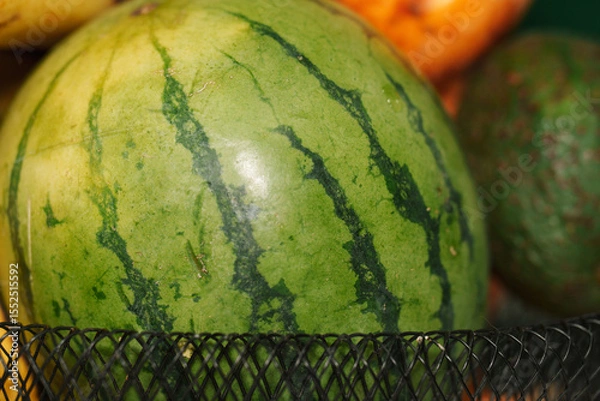 Fototapeta Close-up of a ripe watermelon resting in a black mesh fruit basket, surrounded by other fruits