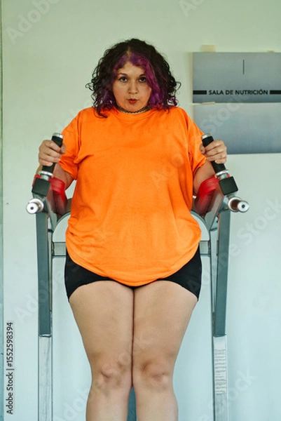 Fototapeta Beautiful overweight Latina woman wearing colorful 80s-style clothing and makeup, exercising with abdominal muscles on a machine in a gym, very positive. Effort, improvement, and overcoming.
