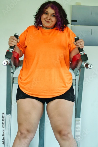 Fototapeta Beautiful overweight Latina woman wearing colorful 80s-style clothing and makeup, exercising with abdominal muscles on a machine in a gym, very positive. Effort, improvement, and overcoming.