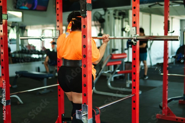 Fototapeta View of an overweight young Latina woman lifting weights and exercising with weights in a gym. Pushing herself. Improvement and well-being.