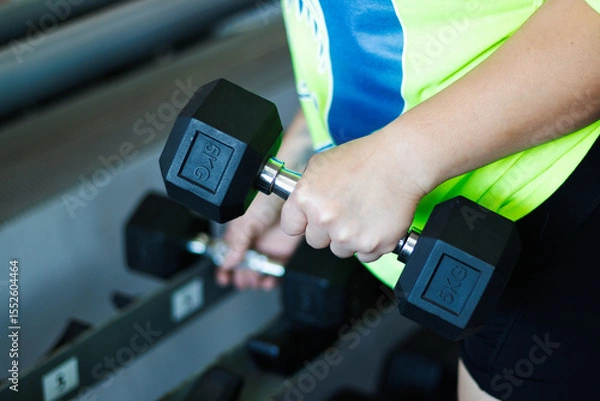 Fototapeta Arm view of a young, overweight Latina woman exercising with weights in a gym, pushing herself, and looking at weights. Health, improvement, and well-being.