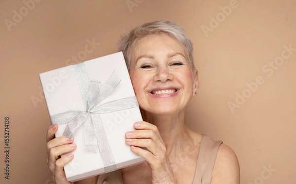 Obraz Portrait of cheerful positive glad charming aged woman having gift box over beige backgrou