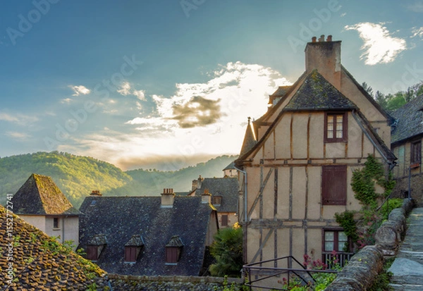 Fototapeta Panoramic view of the historic village of Conques with rustic medieval houses with the mountains in the background illuminated by the golden light of sunset under a sky with backlit clouds.