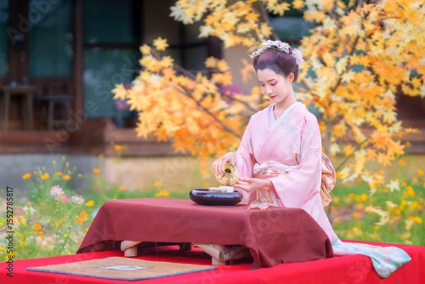 Fototapeta Asian woman wearing a traditional Furisode Kimono performs a tea ceremony happily beside a colorful, vibrant maple tree in a garden with the autumn colors.