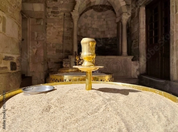 Fototapeta Photo of a brass candle holder in sand-filled tray at the entrance of Panagia Ekatontapiliani church in Parikia, Paros Island, framed by Byzantine arches and historic stonework.