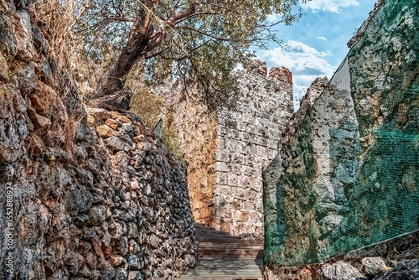 Fototapeta Stone path leads through the ancient walls of Alanya Kalesi, surrounded by rocky structures and greenery, Turkey. Historic castle weathered stones contrast on the bright sky and vibrant vegetation