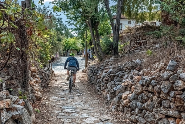 Fototapeta A cyclist rides along a rustic stone path amid trees and ancient walls at Alanya Castle. The peaceful setting highlights the site's scenic, historic charm. Alanya, Turkey - October 24, 2020